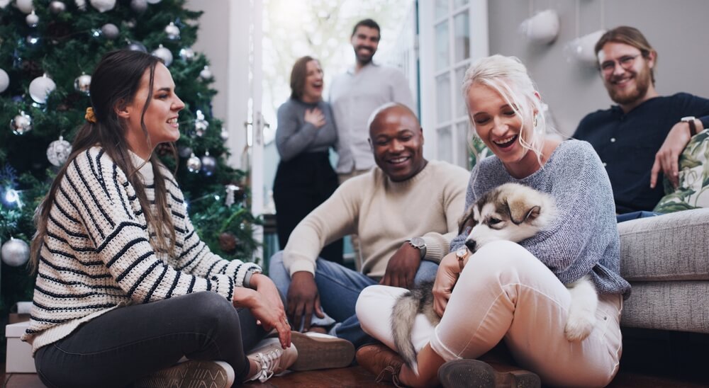Group of friends laughing with a Husky puppy by a Christmas tree, representing pet holiday safety in San Diego